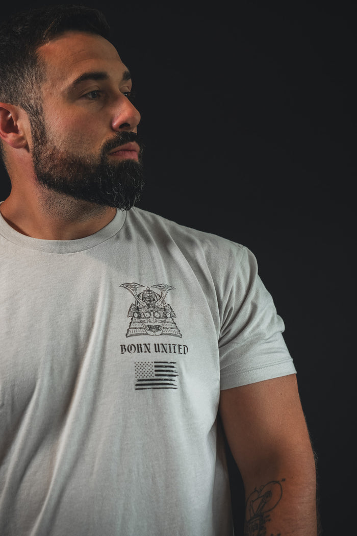 A bearded man with short dark hair wears the "With Honor" tee by Born United Private Label—light gray with a bold graphic, standing against a dark backdrop as a modern warrior symbolizing honor and integrity.