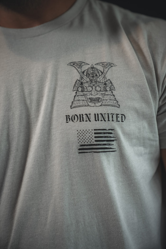 A person wearing the With Honor light gray T-shirt by Born United Private Label, featuring a samurai helmet, BORN UNITED text, and a stylized American flag—ideal for those who value honor and unity.