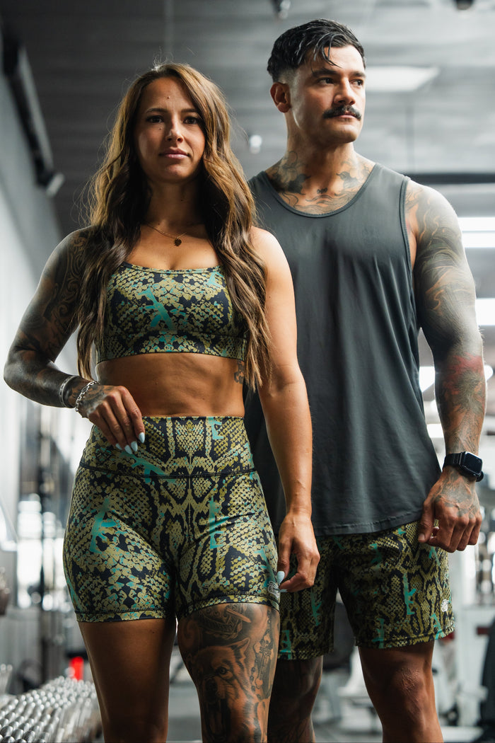 A woman and man with visible tattoos and athletic builds wear matching Tango Charlie Apparel Lethal Venom outfits, including Women's High-Waisted Seamless Front Compression Training Shorts, posing confidently in a gym with weights and equipment.