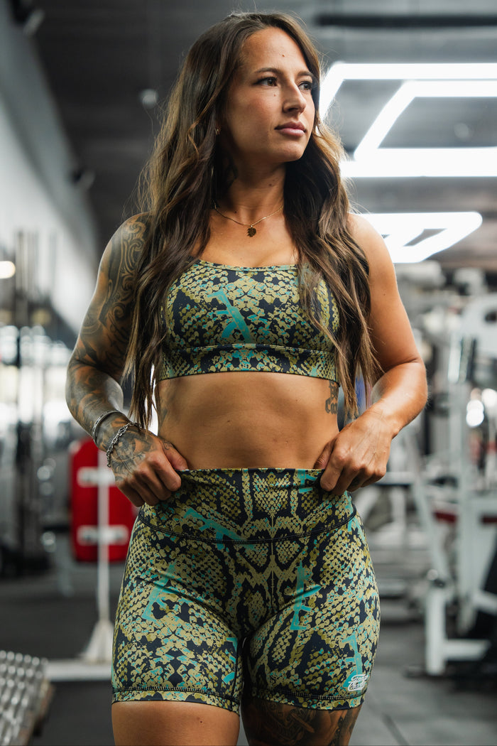 A woman models Tango Charlie Apparel's Lethal Venom Women's High-Waisted Seamless Front Compression Training Shorts, standing confidently in a gym with her hands on her hips and gym equipment visible in the background.
