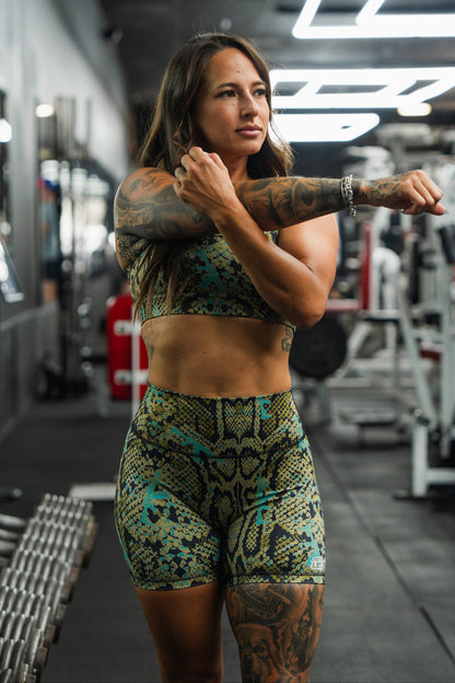 A woman wearing Tango Charlie Apparel's Women's High-Waisted Seamless Front Compression Training Shorts in Lethal Venom stretches in a gym, surrounded by equipment and dumbbells, focused on her workout.