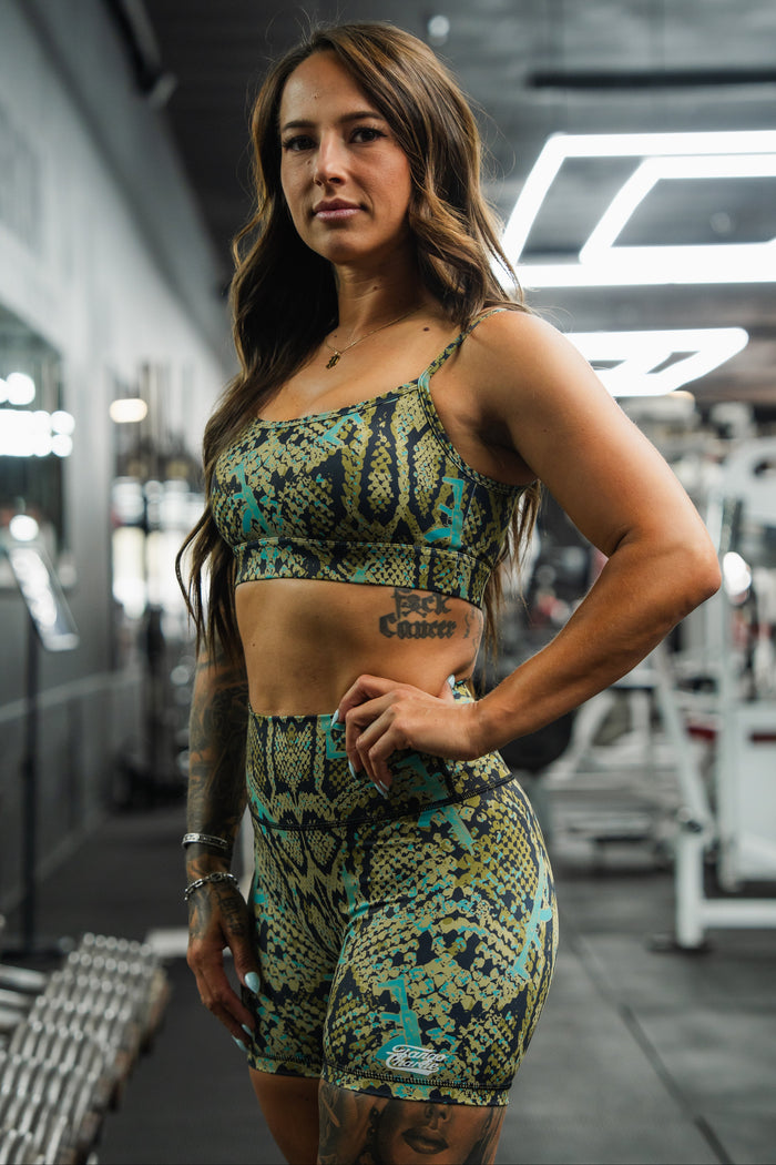 A woman wearing Tango Charlie Apparel's Women's High-Waisted Seamless Front Compression Training Shorts in Lethal Venom stands confidently in a gym, surrounded by fitness equipment, with her hand on her hip.