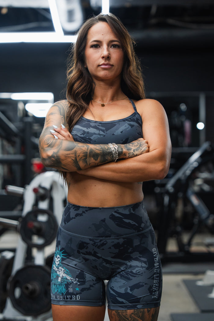 A confident, tattooed woman with long brown hair stands in a gym wearing Born United Private Label's Women’s High-Waisted Seamless Front Compression Training Shorts | Liberty or Death, paired with a matching sports bra amid exercise equipment.