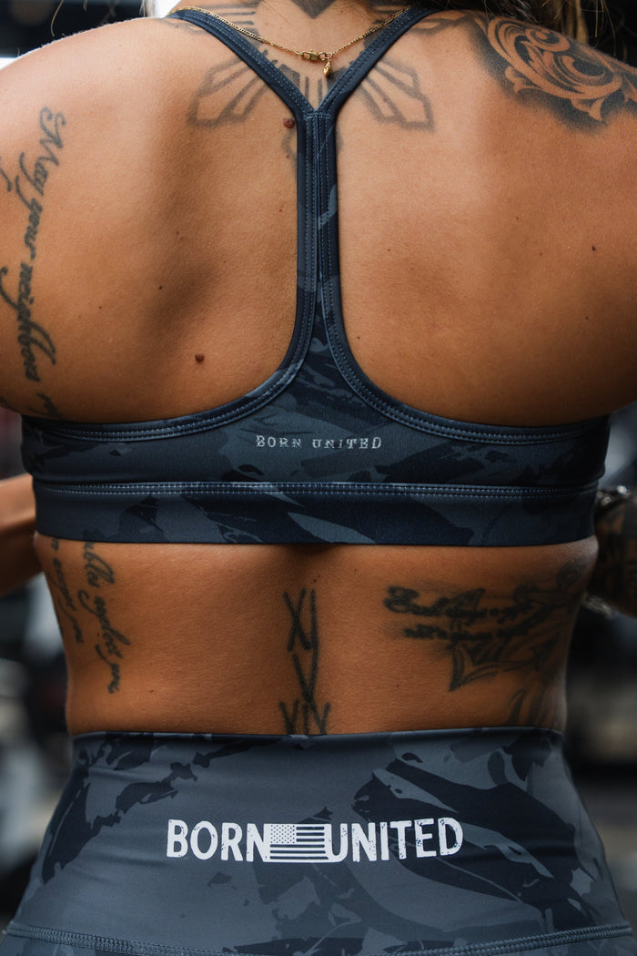 A tattooed person wears a matching dark sports bra and Born United Private Label Women's High-Waisted Seamless Front Compression Training Shorts (Liberty or Death), with focus on their upper back and shoulders.