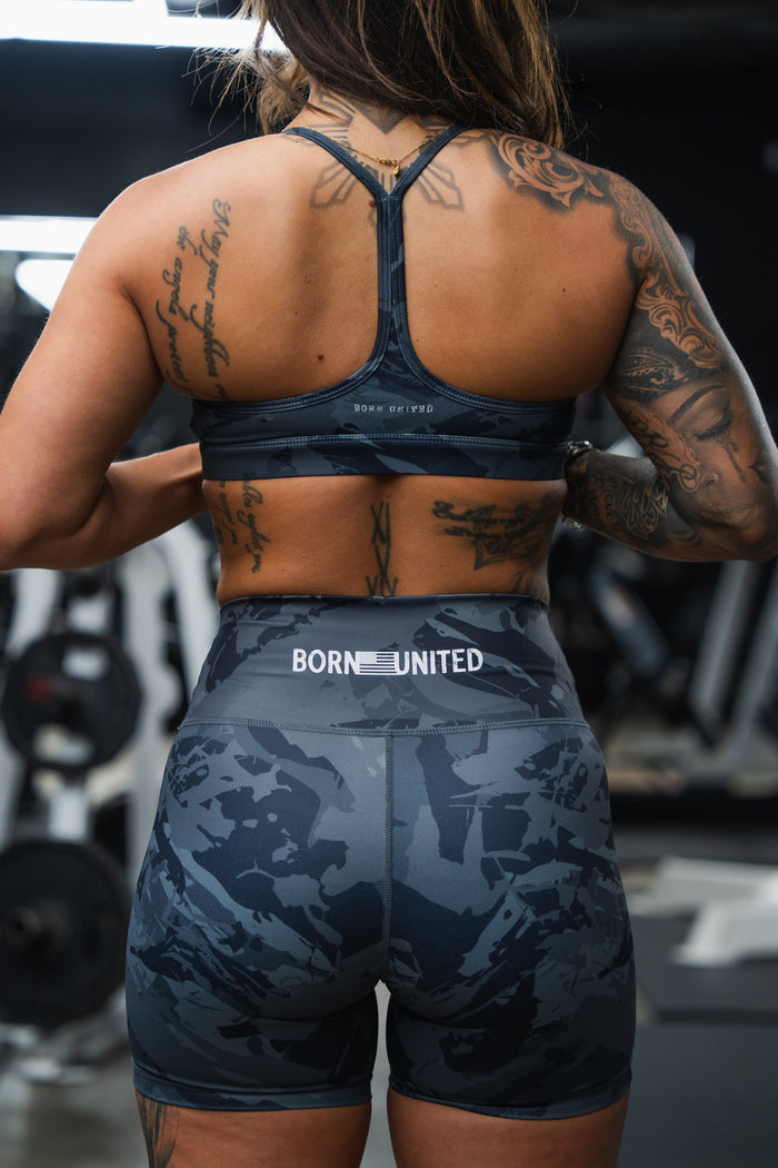 A tattooed woman in a blue sports bra and Born United Private Label Women's High-Waisted Seamless Front Compression Training Shorts (Liberty or Death) stands in a gym with "BORN UNITED" on the waistband, gym equipment behind her.