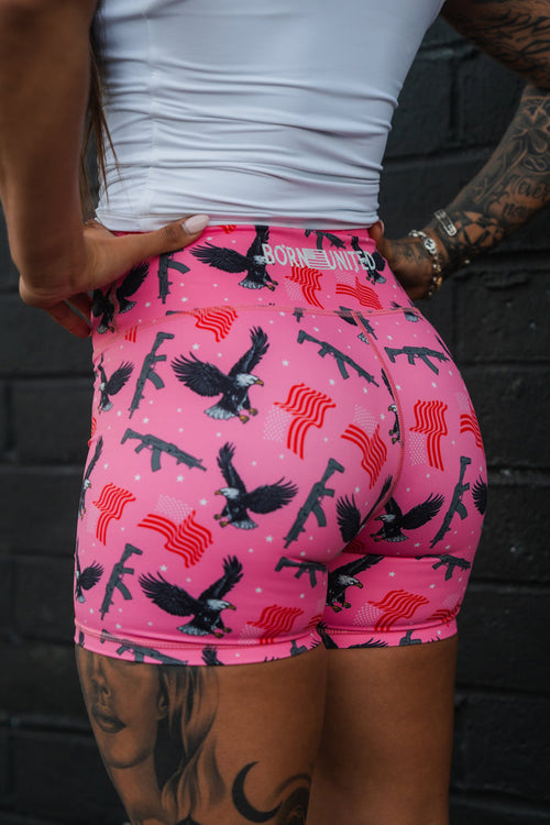 A tattooed person in a white top and Born United Private Labels Womens High-Waisted Seamless Front Compression Training Shorts in Pink Victory, featuring eagles, rifles, and red stripes on breathable fabric, poses confidently with hands on hips against a black brick wall.