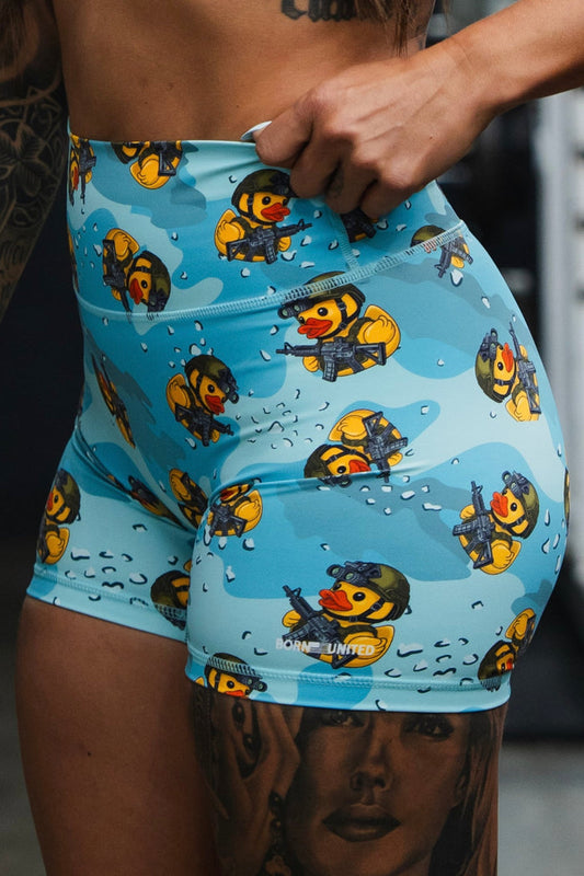 A person with arm and leg tattoos wears Born United Private Label's TactiQuack Women's High-Waisted Seamless Front Compression Training Shorts in blue, featuring cartoon soldier rubber ducks, while holding the waistband.
