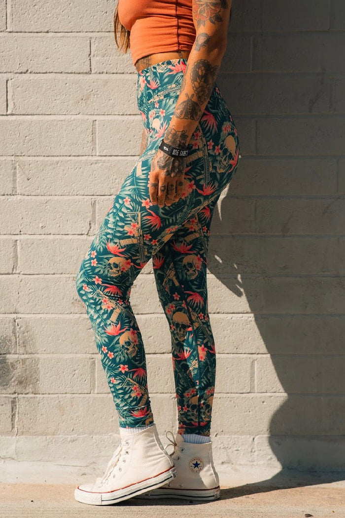 A tattooed person in a sleeveless orange top, Tango Charlie Apparel Women's Performance Leggings | Aloha AR, and white Converse sneakers stands sideways against a sunlit light brick wall.