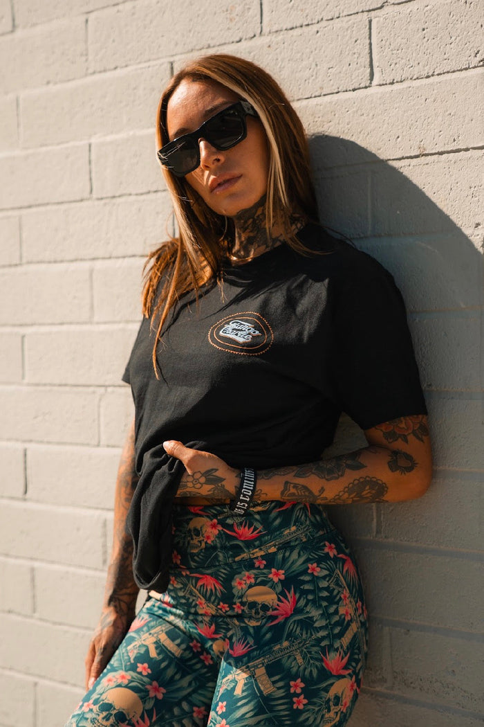 A woman with long hair and tattoos wears sunglasses, a black t-shirt, and Tango Charlie Apparel's Women's Performance Leggings | Aloha AR, standing confidently against a light brick wall in bright sunlight.