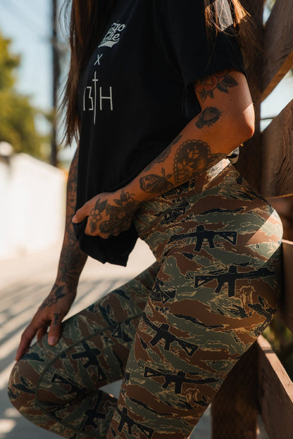 A person with tattooed arms wears a black t-shirt and Tango Charlie Apparel Women's Performance Leggings | Snipes & Stripes, featuring a high-rise fit and firearm print, standing outdoors beside a wooden fence in sunlight.