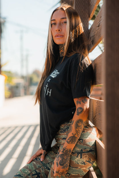 A woman with long brown hair and arm tattoos poses confidently outdoors in bright sunlight, wearing Tango Charlie Apparel's Women's Performance Leggings | Snipes & Stripes and a black t-shirt, leaning against a wooden fence.