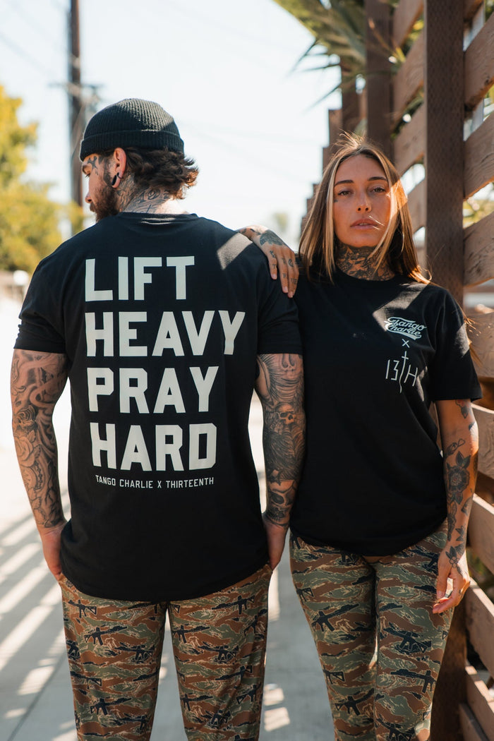 Two tattooed people in camo pants and black tees stand side by side outdoors. The woman wears Tango Charlie Apparel's Women's Performance Leggings | Snipes & Stripes with a high rise tee, while the man's shirt reads LIFT HEAVY PRAY HARD.
