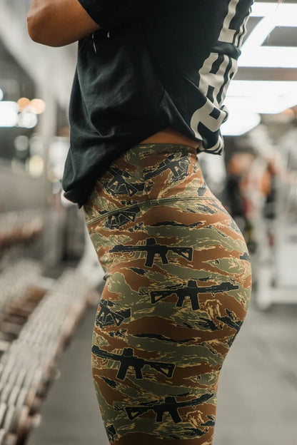 A woman in Tango Charlie Apparel's Women's Performance Leggings | Snipes & Stripes, featuring tummy control and black rifle motifs, stands sideways in a bright gym beside weights and equipment, showcasing the leggings' sleek design.