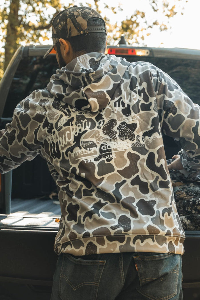 A person wearing the Mucker Bucket Wood Ducks Premium Athletic Duck Camo Hoodie stands at an open pickup tailgate, loading gear into the truck bed with trees and sunlight in the background.