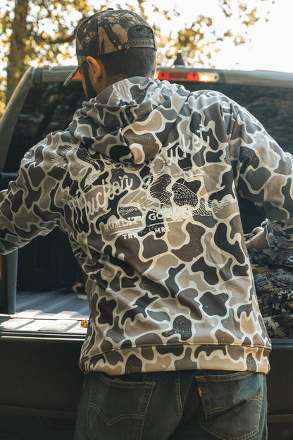 A person wearing the Mucker Bucket Wood Ducks Premium Athletic Duck Camo Hoodie stands at an open pickup tailgate, loading gear into the truck bed with trees and sunlight in the background.