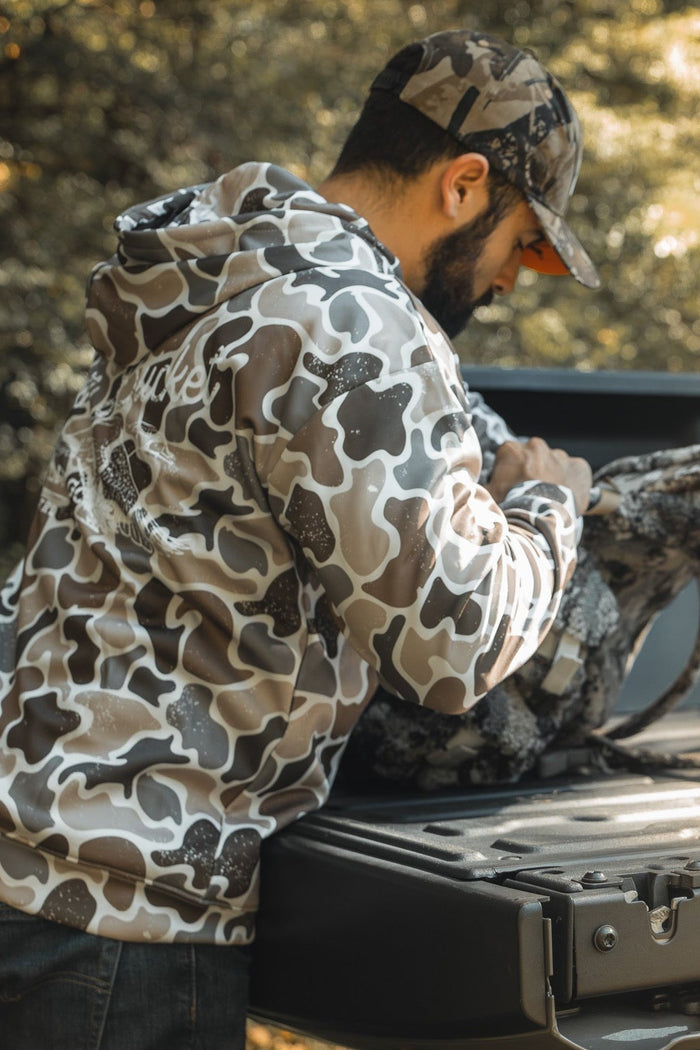 A man in a Mucker Bucket Wood Ducks Premium Athletic Duck Camo Hoodie and cap stands at a pickup truck's open tailgate, looking into a camo backpack as sunlight filters through the trees in the background.