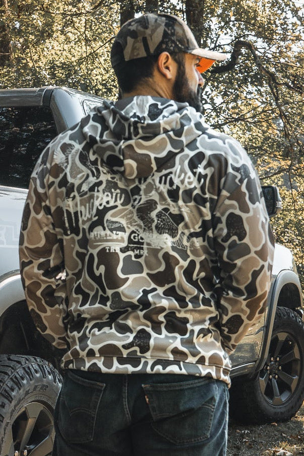 A man in a Mucker Bucket Wood Ducks Premium Athletic Duck Camo Hoodie and cap stands by a silver truck in a sunlit, wooded area, showcasing the hoodie’s quality in a natural outdoor setting.