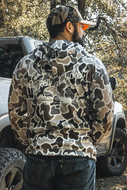 A man in a Mucker Bucket Wood Ducks Premium Athletic Duck Camo Hoodie and cap stands by a silver truck in a sunlit, wooded area, showcasing the hoodie’s quality in a natural outdoor setting.