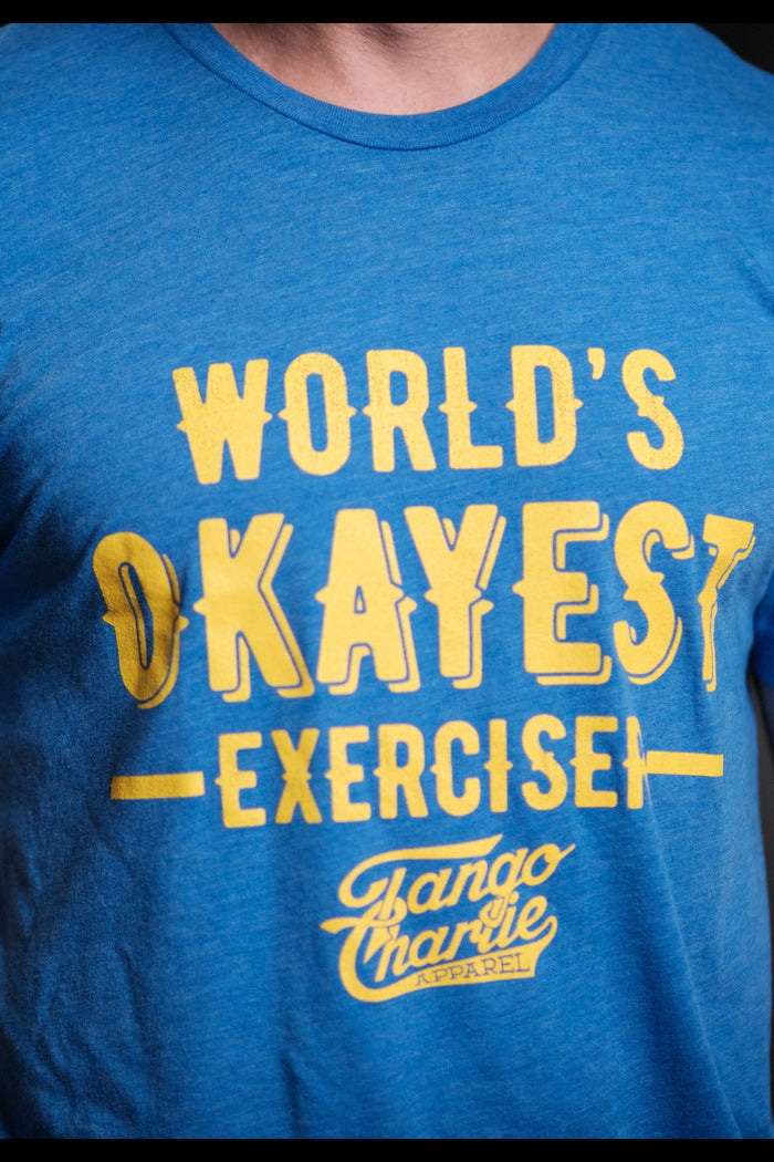 Wearing a comfy cotton/polyester blend tee, a person showcases the blue Worlds OKAYEST Exerciser T-shirt by Tango Charlie Apparel, complemented by tattoos on their right arm for an edgy look.