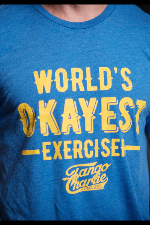 Wearing a comfy cotton/polyester blend tee, a person showcases the blue Worlds OKAYEST Exerciser T-shirt by Tango Charlie Apparel, complemented by tattoos on their right arm for an edgy look.