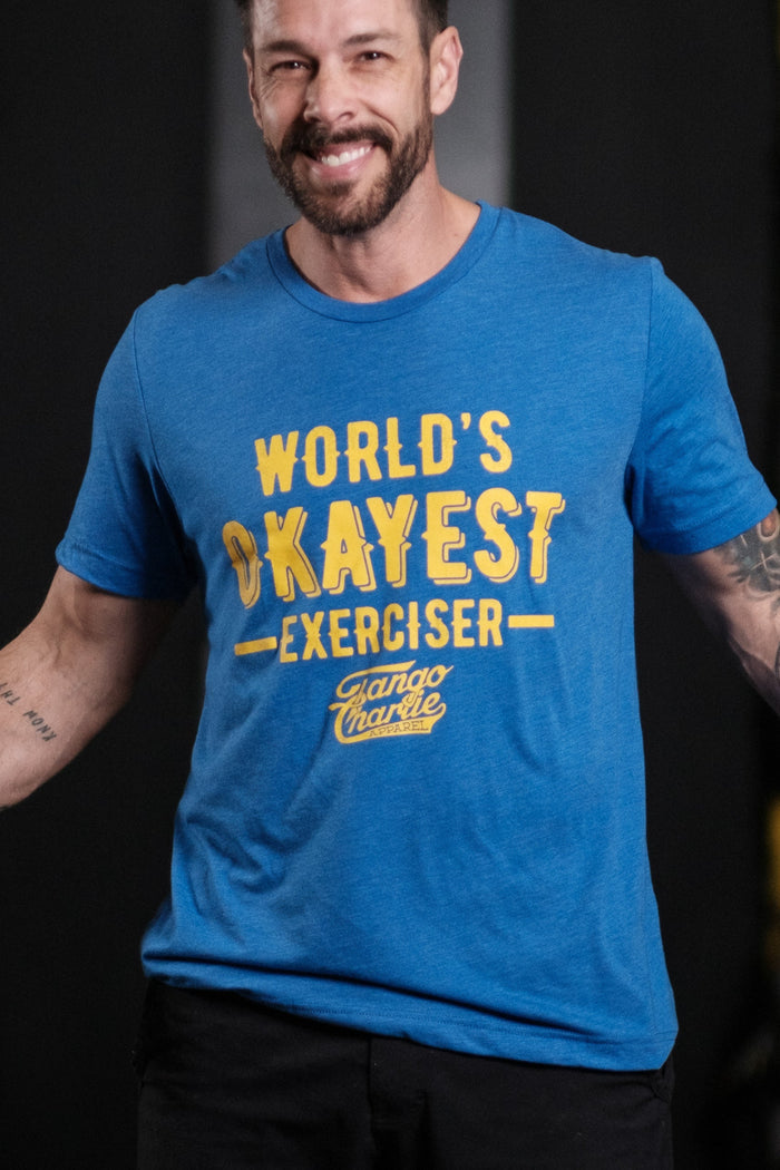 A bearded man stands smiling with arms outstretched, wearing a comfy Worlds OKAYEST Exerciser Tee by Tango Charlie Apparel. The cotton/poly blend shirt features bold yellow text against a blurred gym backdrop, creating an amusing scene.