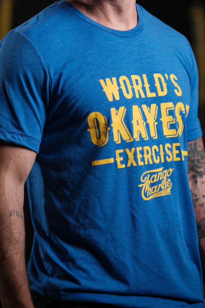 A person dons the Tango Charlie Apparels Worlds OKAYEST Exerciser Tee, a comfy blue cotton/poly blend with yellow text. Their tattooed arm adds flair against a dark, blurred background.