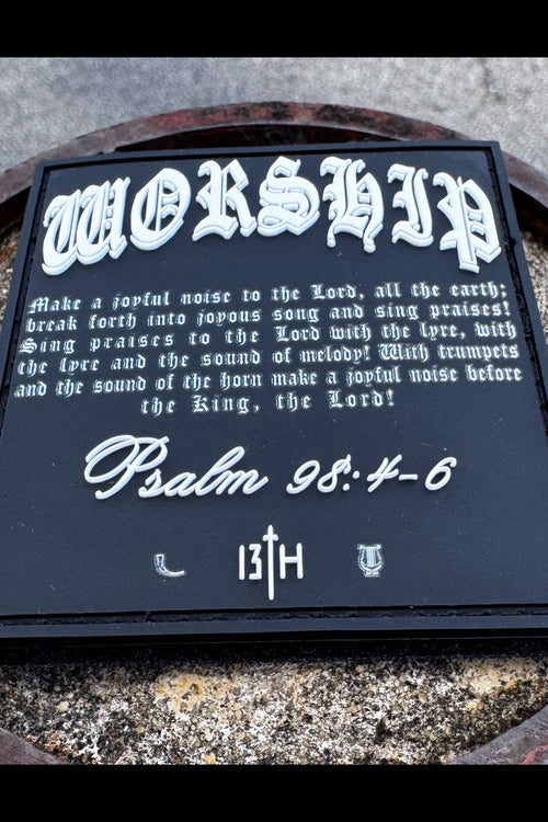 The Worship PVC Patch by 13th features Psalm 98:4-6 in ornate white text on black, with 13TH and two small symbols at the bottom, shown resting on a stone surface.