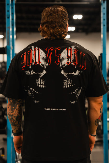 A man with tattooed arms stands in a gym wearing the Tango Charlie Apparel You vs You Premium Oversized Tee, featuring two skulls and bold red YOU VS YOU text. Gym equipment and weights are visible in the background.