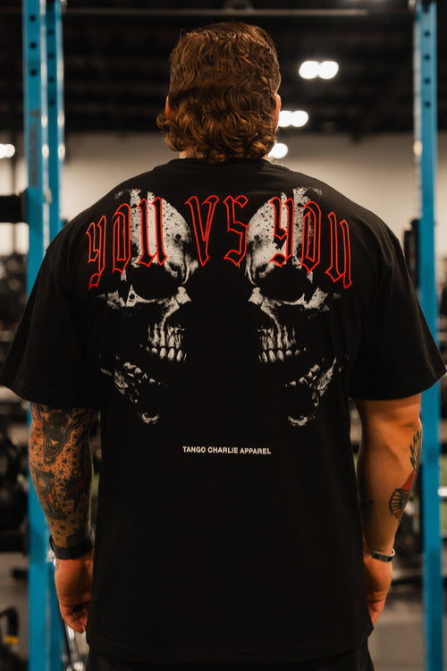 A man with tattooed arms stands in a gym wearing the Tango Charlie Apparel You vs You Premium Oversized Tee, featuring two skulls and bold red YOU VS YOU text. Gym equipment and weights are visible in the background.