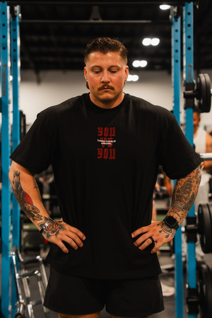 A tattooed man stands in a gym, hands on hips, wearing the You vs You - Premium Oversized Tee from Tango Charlie Apparel. Weightlifting equipment and other people are visible in the background.