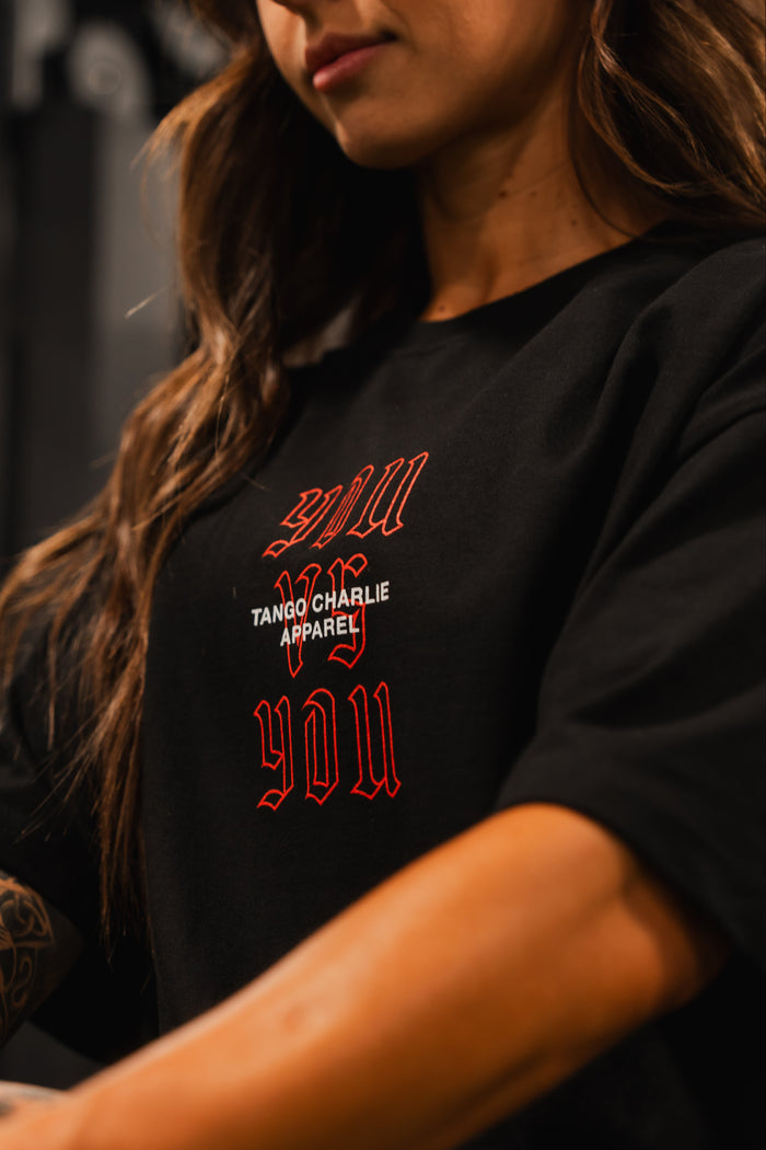 A woman with long brown hair and a tattooed arm wears the You vs You - Premium Oversized Tee by Tango Charlie Apparel. The focus is on her upper body and shirt, highlighting the tees premium heavyweight cotton quality.