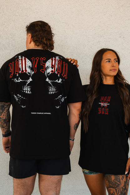 A man and woman wear Tango Charlie Apparel’s You vs You - Premium Oversized Tee. The man displays the skull back graphic, while the woman faces forward, showing the unisex tee’s front design against a plain wall.