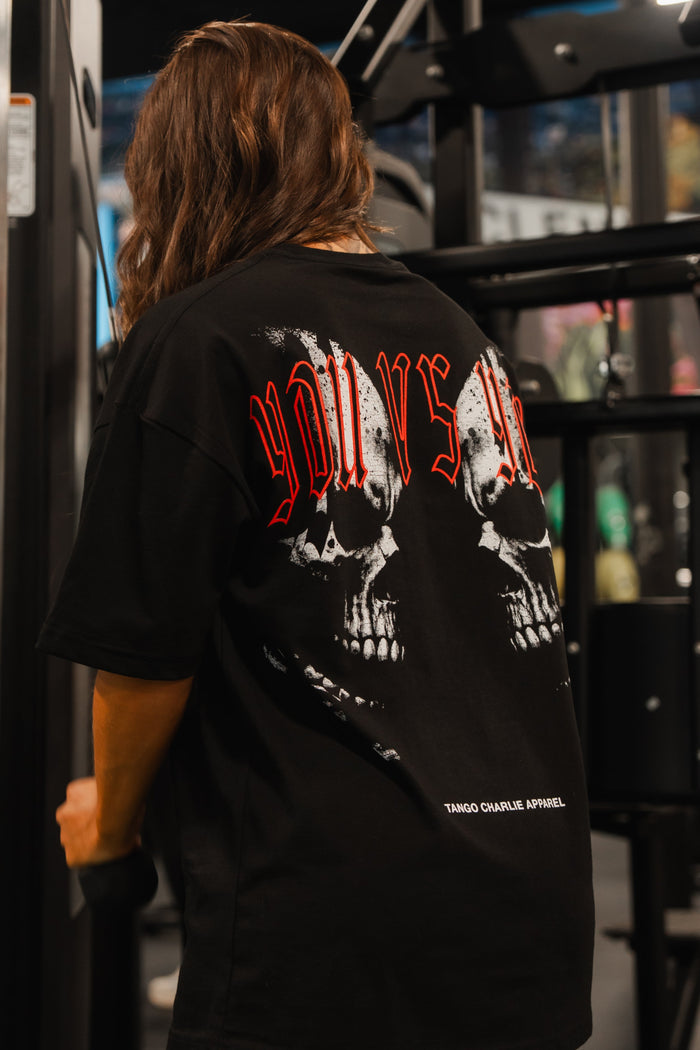 A person with long brown hair and tattooed legs stands in a gym, back to the camera, wearing the Tango Charlie Apparel You vs You Premium Oversized Tee featuring skull graphics and red lettering.