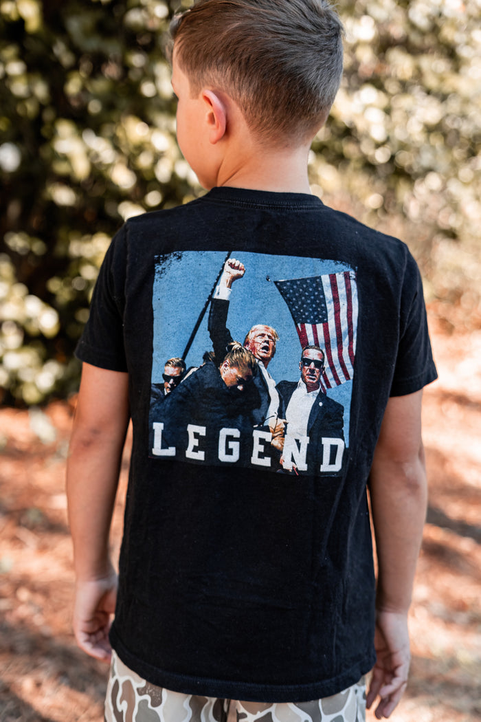 A boy in a Youth Forever Legend tee from Born United Private Label, featuring four men with an American flag, stands outside against a backdrop of trees, exuding subtle patriot pride.