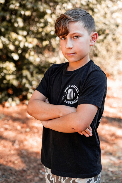 A young boy with short, styled hair stands confidently with arms crossed, wearing a Youth Forever Legend tee by Born United Private Label. His shorts are patterned. The background features blurred trees and sunlight, suggesting a natural setting.