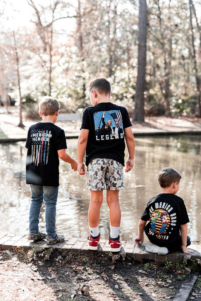 By a pond, three boys stand. The left boy wears a Merica Hero tee, exuding patriot vibes. The center boy sports the Born United Private Label Youth Forever Legend shirt and camo shorts, while the right boy, seated, showcases his Hero Member shirt. Trees reflect in the water behind them.